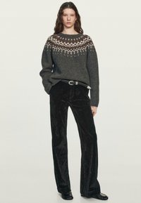 Grauer Wollpullover mit gemustertem Einsatz in Creme und Braun, kombiniert mit schwarzen Samt-Hose mit ausgestelltem Bein. Modell steht selbstbewusst.