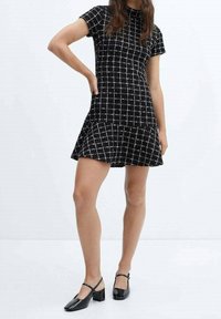 Mujer vestido de cuadros en blanco y negro de manga corta con un dobladillo acampanado y zapatos de tacón bloque negros estilo Mary Jane, posando con la mano en la cadera.