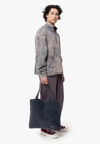 Pullover oversized grigio-marrone con mezza zip, dotato di tasca frontale. Il modello porta una borsa a spalla in pelliccia sintetica blu navy con doppi manici.