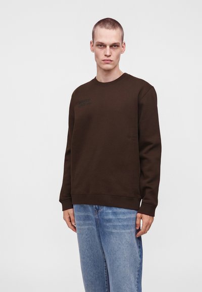 Jeune homme aux cheveux courts portant un pull marron foncé à manches longues et un jean bleu clair, debout devant un fond clair uni.