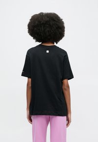 Μαύρο κοντομάνικο μπλουζάκι από βαμβάκι, με στρογγυλή λαιμόκοψη και μικρή ετικέτα με λογότυπο στο πάνω μέρος της πλάτης. Φοριέται με ροζ παντελόνια.