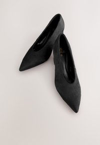 Par svart spetsiga högklackade skor i mjukt, texturerat material på en ljus neutral bakgrund, vinklade med klackarna inåt.