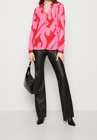 Blusa rossa e rosa a fantasia con scollatura a V, maniche lunghe e chiusura a bottoni, abbinata a pantaloni a zampa d'elefante in ecopelle nera e una borsa nera.