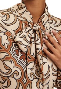 Mönstrad blus i beige med intrikata bruna och svarta mönster. Har en knutkrage och en mjuk tygstruktur.