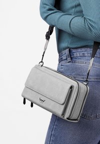 Osoba v modrém žebrovaném svetru a džínech nese texturovanou světle šedou krosnu s černým nastavitelným popruhem a zipovými kapsami.