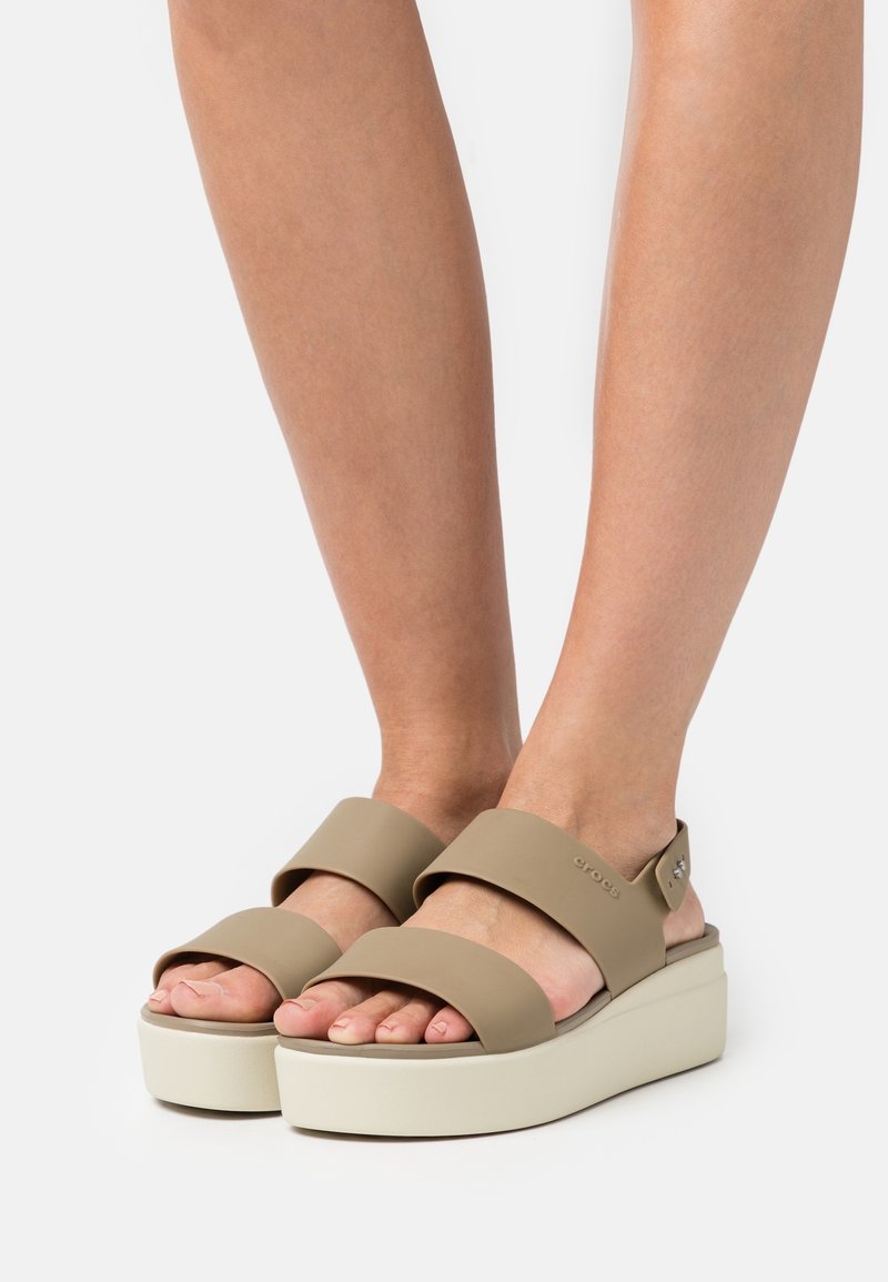 Pieds portant des sandales compensées beiges avec deux larges brides sur les orteils et le dessus du pied sur fond blanc.