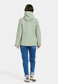 Vaaleanvihreä sadetakki hupulla, jossa on sileä pinta ja säädettävät hihansuut, yhdistettynä sinisiin farkkuihin ja valkoisiin tennareihin.