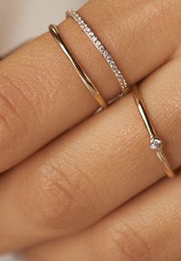 Tre anelli d'oro impilati sulle dita: un band liscia, uno decorato con piccole pietre trasparenti e uno con un singolo accento di pietra.