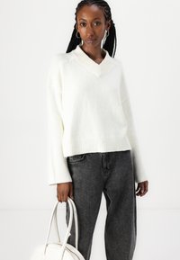 Hvit strikket genser med V-hals og ribbet kant, sammen med høyreiste mørke grå jeans. Modellen holder en hvit håndveske.