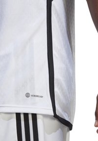 Weißes Sporttrikot mit strukturiertem Muster, schwarzen, gesäumten Kanten und "AEROREADY"-Logo in der Nähe des Saums. Glattes, leichtes Material.