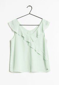 Blusa sin mangas de color verde claro con escote en V y detalle de volantes, colgada en una percha negra sobre un fondo blanco.