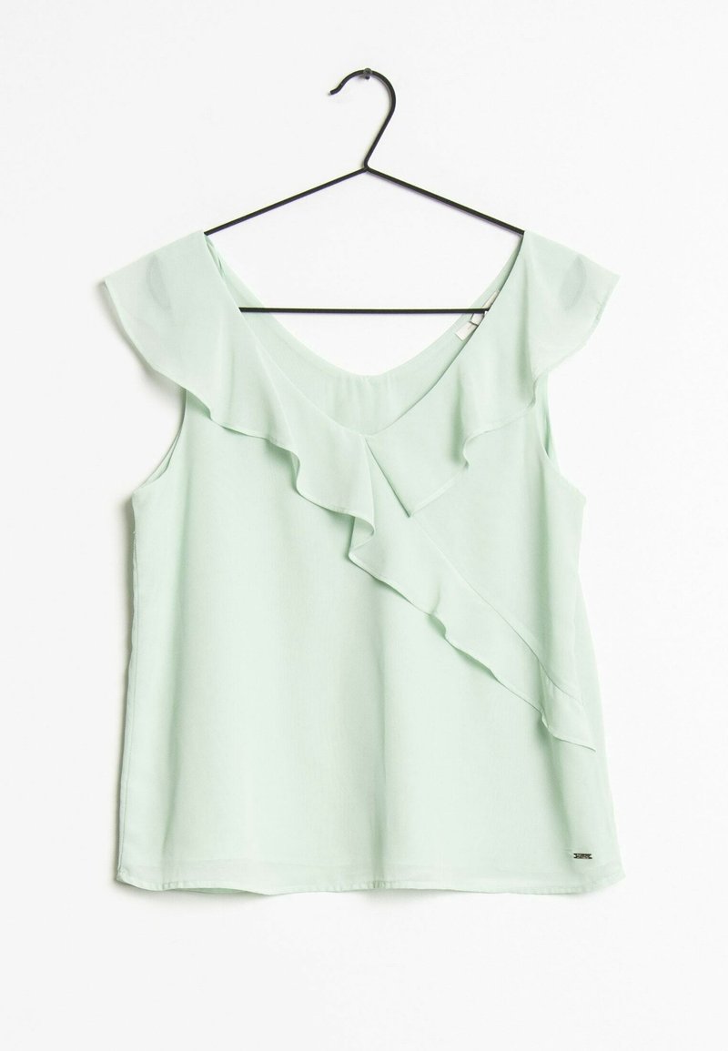 Blusa sin mangas de color verde claro con escote en V y detalle de volantes, colgada en una percha negra sobre un fondo blanco.