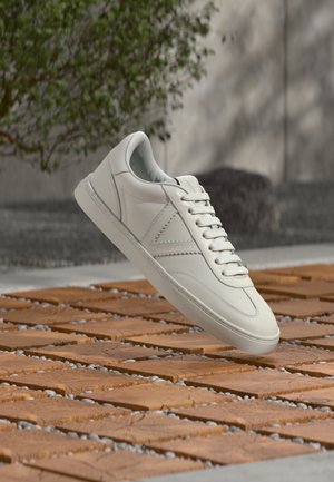Zapatilla deportiva de cuero blanco con cordones flotando sobre un bloque de madera y superficie exterior de guijarros, planta verde y piedra desenfocadas al fondo.