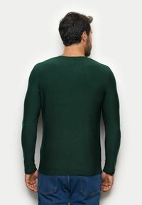 Zielona koszula z długim rękawem, okrągłym dekoltem, dopasowanym krojem, gładką fakturą i minimalną liczbą szwów. Noszona z niebieskimi dżinsami.