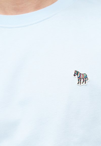 Ανοιχτό μπλε μπλουζάκι με μικρό κεντημένο πατς ζέβρας με πολύχρωμες ρίγες στην αριστερή πλευρά του στήθους.