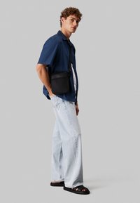 Borsa a tracolla nera, camicia leggera a maniche corte blu, canotta bianca, jeans larghi azzurri e ciabatte nere.