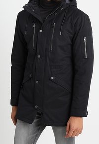 Parka negra con capucha, que presenta bolsillos en el pecho y los lados con cremallera, botones a presión y una tela de textura suave.