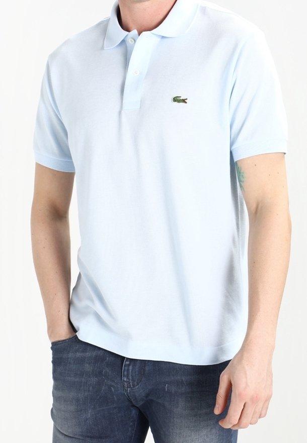 Homme portant un polo bleu clair à manches courtes avec un petit logo crocodile et un jean bleu, main gauche dans la poche, debout.