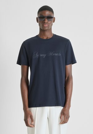 Modello maschile che indossa una T-shirt Antony Morato blu scuro, pantaloni bianchi e grandi occhiali da sole scuri, in piedi davanti a uno sfondo chiaro e semplice.