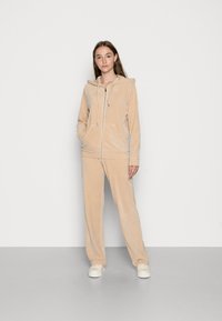 Beige velour-huppari, jossa on vetoketju ja kenguruhyppypusseja sek�ä vastaavat löysät housut. Tasainen tekstuuri ilman kuvioita.