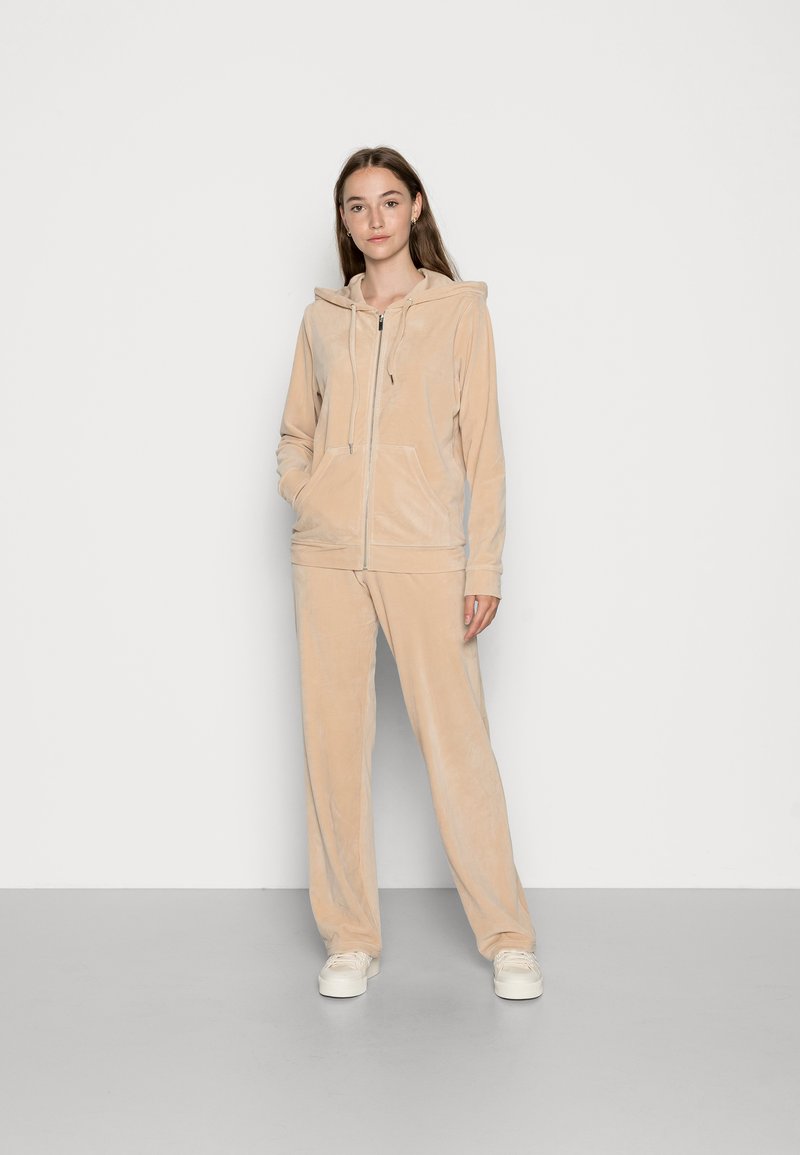 Beige Velour-Trainingsanzug mit einer Kapuzenjacke mit Reißverschluss und Kängurutaschen sowie passenden, locker sitzenden Hosen. Glatte Textur ohne Muster.