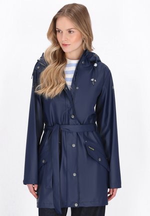 Donna che indossa un impermeabile blu navy con cintura e cappuccio sopra una camicia bianca a righe blu, in piedi davanti a uno sfondo bianco semplice.