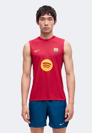 Jeune homme portant un maillot de football rouge sans manches du FC Barcelone avec le logo jaune Spotify et un short Nike bleu, debout devant un fond uni.