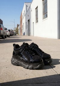 Para czarnych masywnych sneakersów z teksturowanymi i błyszczącymi panelami na słonecznym, miejskim chodniku w pobliżu zaparkowanych samochodów i białych budynków.