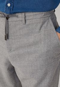 Primo piano di una persona che indossa pantaloni grigi con coulisse e una camicia button-up blu, con una mano in tasca.