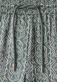 Pantalones cortos verdes con un patrón geométrico blanco, que cuentan con una cinturilla elástica, cordón con extremos metálicos plateados y una tela suave y texturizada.