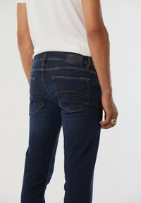 Jean en denim bleu foncé avec une coupe ajustée, comportant des coutures et des accents de poches uniques. Étiquette en cuir noir au niveau de la taille.