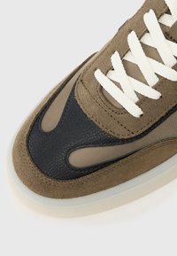 Smeđi i crni tenisice koje kombiniraju nubuk i mrežaste materijale, s bijelim vezicama i glatkim gumenim đonom, istaknute jedinstvenim teksturiranim naglascima.