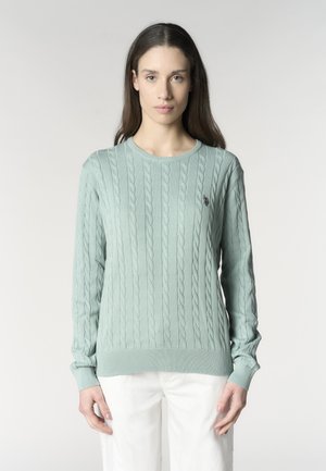 Donna con un maglione azzurro chiaro a trecce e pantaloni bianchi, in piedi davanti a uno sfondo grigio chiaro uniforme.