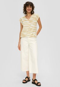 Hellbeige Oberteil mit einem weißen Zebra-Muster, V-Ausschnitt, Kurzarm; kombiniert mit weit geschnittenen weißen Hosen und schwarzen klobigen Sandalen.