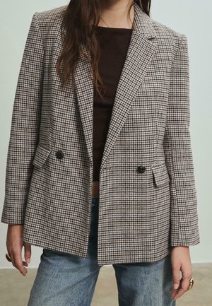 Woman wearing a brown and white houndstooth blazer over a black top and blue jeans, standing with hands relaxed at sides.