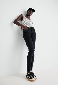 Jeans ajustados negros combinados con una camiseta sin mangas blanca a rayas. Una gorra negra y zapatillas negras con crema completan el atuendo. Diseño sencillo y ajustado.