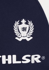 Tessuto blu navy con un emblema ricamato bianco raffigurante uno scudo, foglie di alloro e una "M" stilizzata, insieme a un testo bianco sottostante.