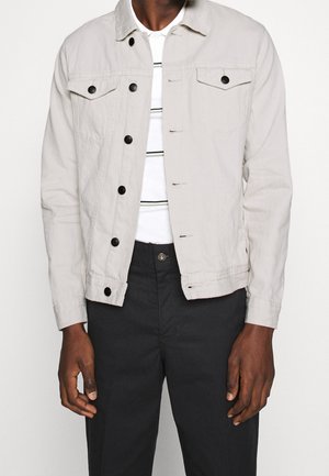 Homme portant une veste beige clair boutonnée par-dessus une chemise blanche à fines rayures noires et un pantalon noir, debout devant un fond uni.