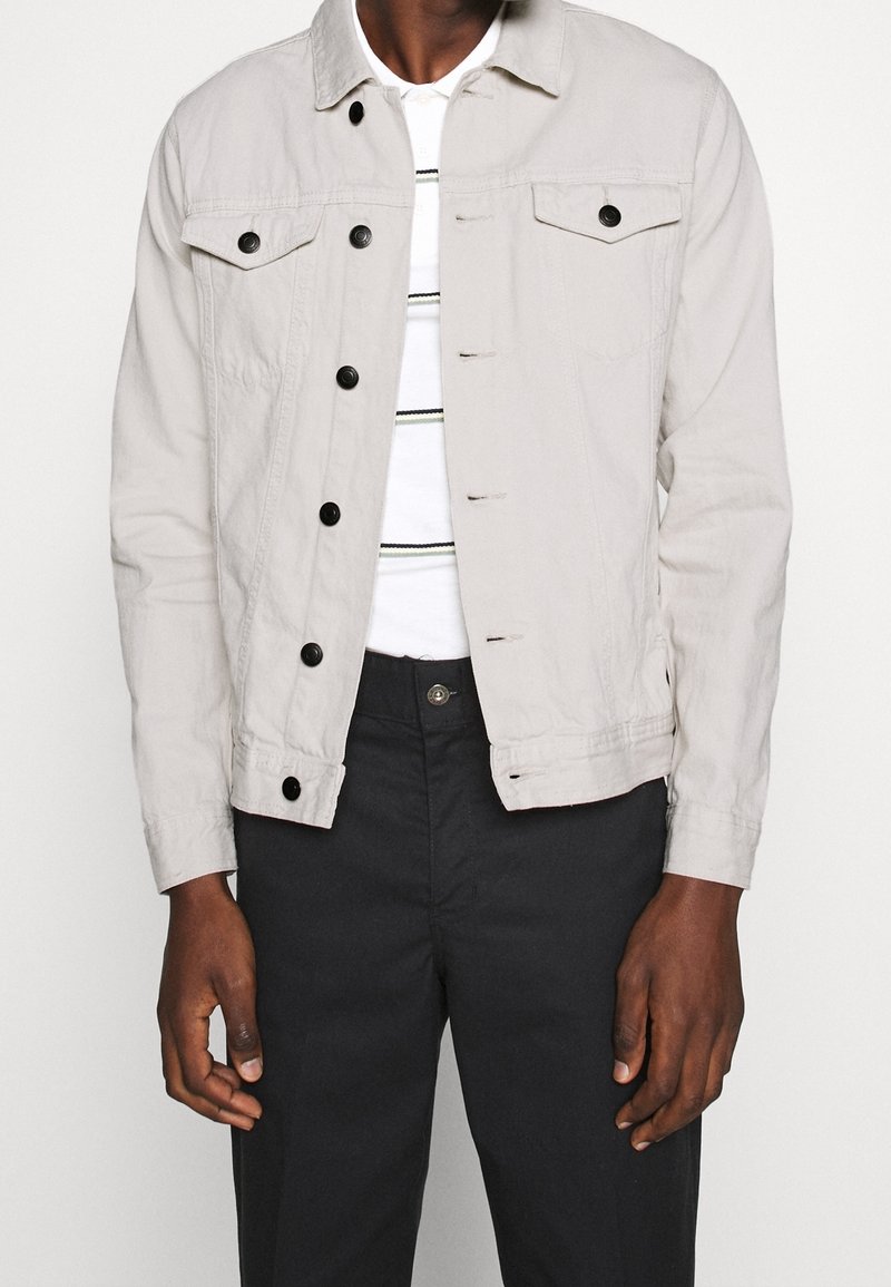 Homme portant une veste beige clair boutonnée par-dessus une chemise blanche à fines rayures noires et un pantalon noir, debout devant un fond uni.