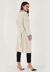 Beige long robe with a textured finish, featuring a patterned collar and wide sleeves, cinched at the waist with a matching belt. Black pants and shoes.