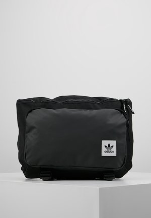 Zwarte Adidas heuptas met voorvak met rits en wit logopatch op een schone witte ondergrond en grijze achtergrond.