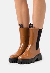 Bottes de pluie aiguille brillantes marron avec une tige en caoutchouc, des panneaux latéraux élastiques noirs et une semelle noire texturée pour une meilleure adhérence.
