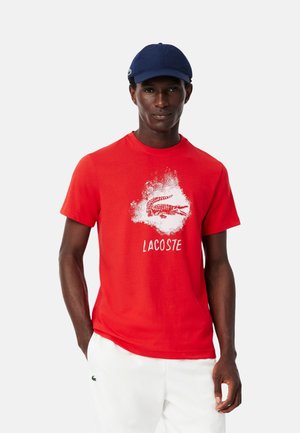 Camiseta de algodón roja con un logo abstracto de cocodrilo blanco y el texto "LACOSTE". Combinada con pantalones blancos y una gorra azul marino.
