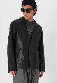 Schwarze Lederblazer mit Reverskragen, zwei vorderen Taschen und Knopfverschluss. Getragen über einem dunkelgrauen Hemd, kombiniert mit Sonnenbrille.
