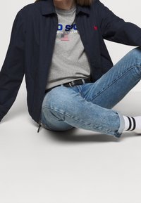 Námořnicky modrá větrovka s límečkem, lehký materiál, červeně vyšívané logo, šedé tričko a modré opotřebené džíny. Černý opasek a proužkované ponožky.