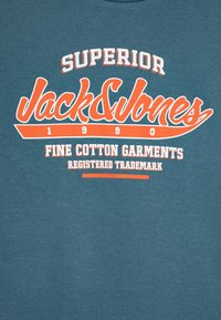Camiseta de algodón en color azul verdoso con texto en naranja brillante: "Superior Jack & Jones", "Prendas de Algodón de Alta Calidad" y "Marca Registrada". Diseño sencillo.