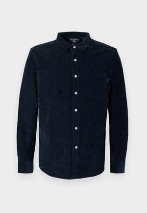 Camisa de pana color marino con mangas largas, cuello abotonado y un bolsillo en el pecho; presenta botones blancos y tejido texturizado.