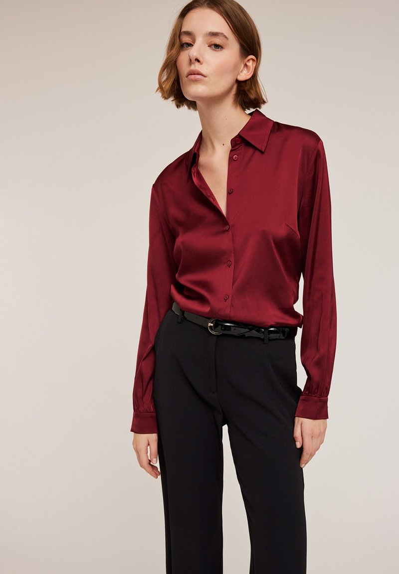 Blouse satin bourgogne à manches longues et design à boutonnage, assortie à un pantalon noir et une ceinture noire, créant une silhouette élégante.