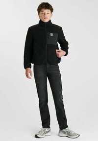 Zwarte fleecejas met hoge kraag, rits aan de voorkant en zak. Gekleed met een donkere spijkerbroek en zilveren sportschoenen. Gladde stof, warme textuur.