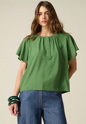 Ung kvinde iført en løs grøn kortærmet top, blå jeans og store grønne og sølvfarvede armbånd på venstre håndled.